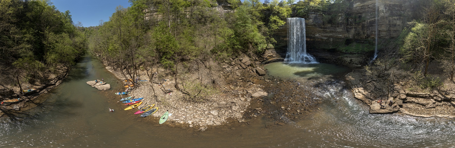 Panoramic photo of waterfalls on the right side with colorful kayaks beached on a rocky shore on the left side.